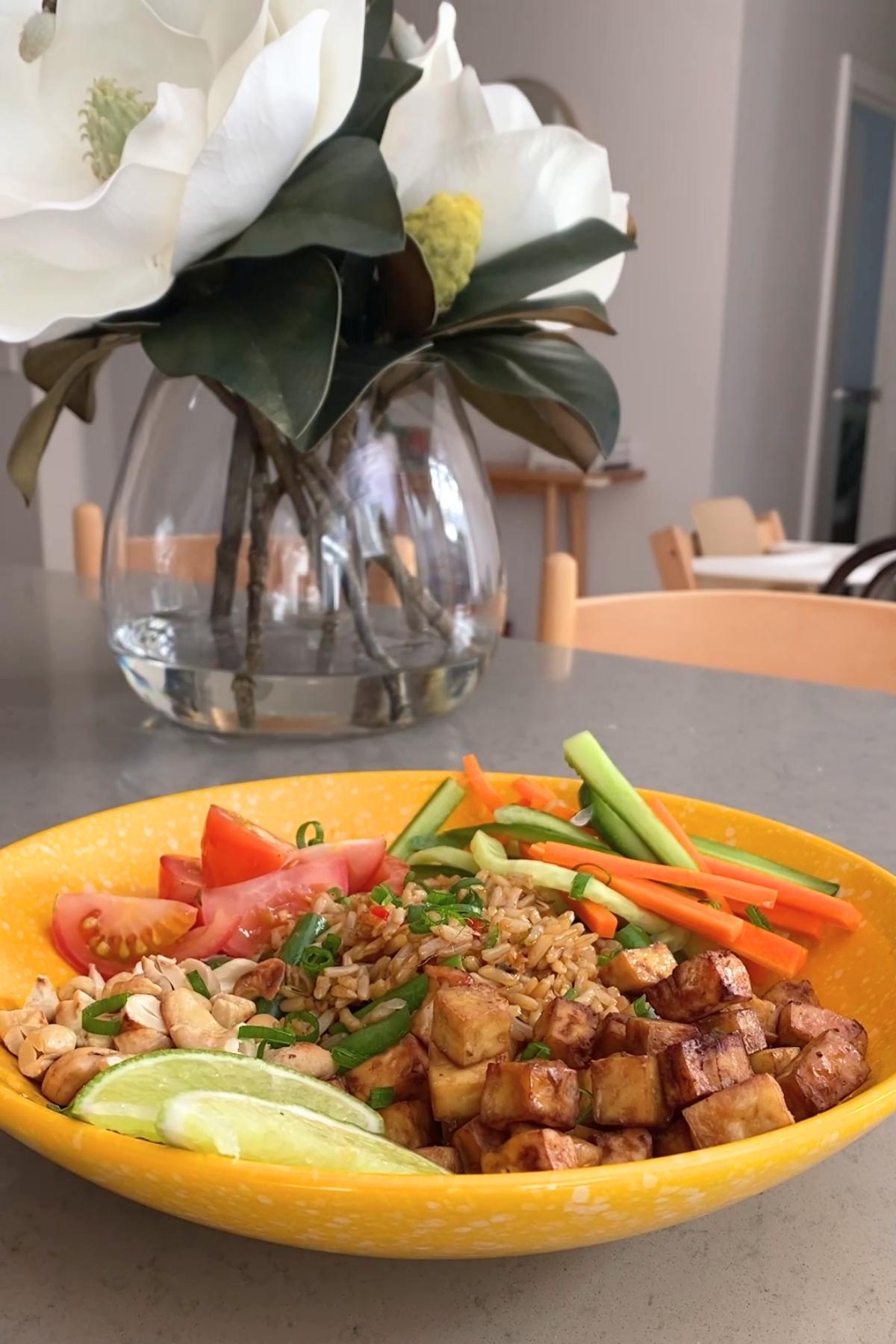 A yellow bowl holding rice, tofu cubes, carrot and cucumber ribbons, tomato wedges, cashews, and lime slices.