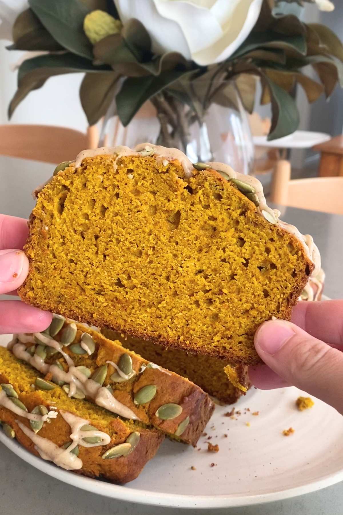 Hand holding slice of pumpkin loaf showing bright orange crumb and pepitas in sunlight.