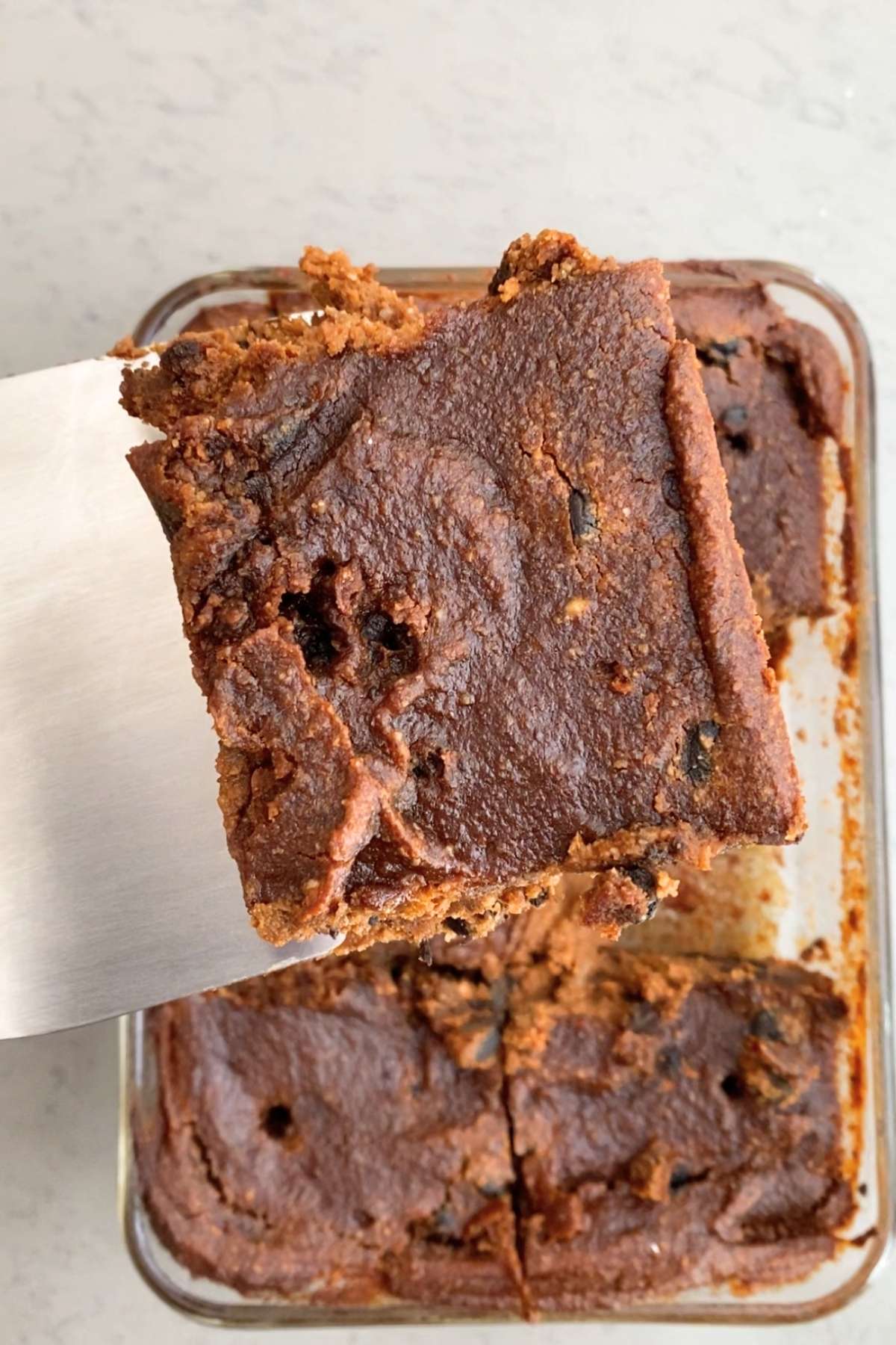A single pumpkin brownie square being lifted from a glass baking dish, showing a dense, crackled top with melted chocolate bits.
