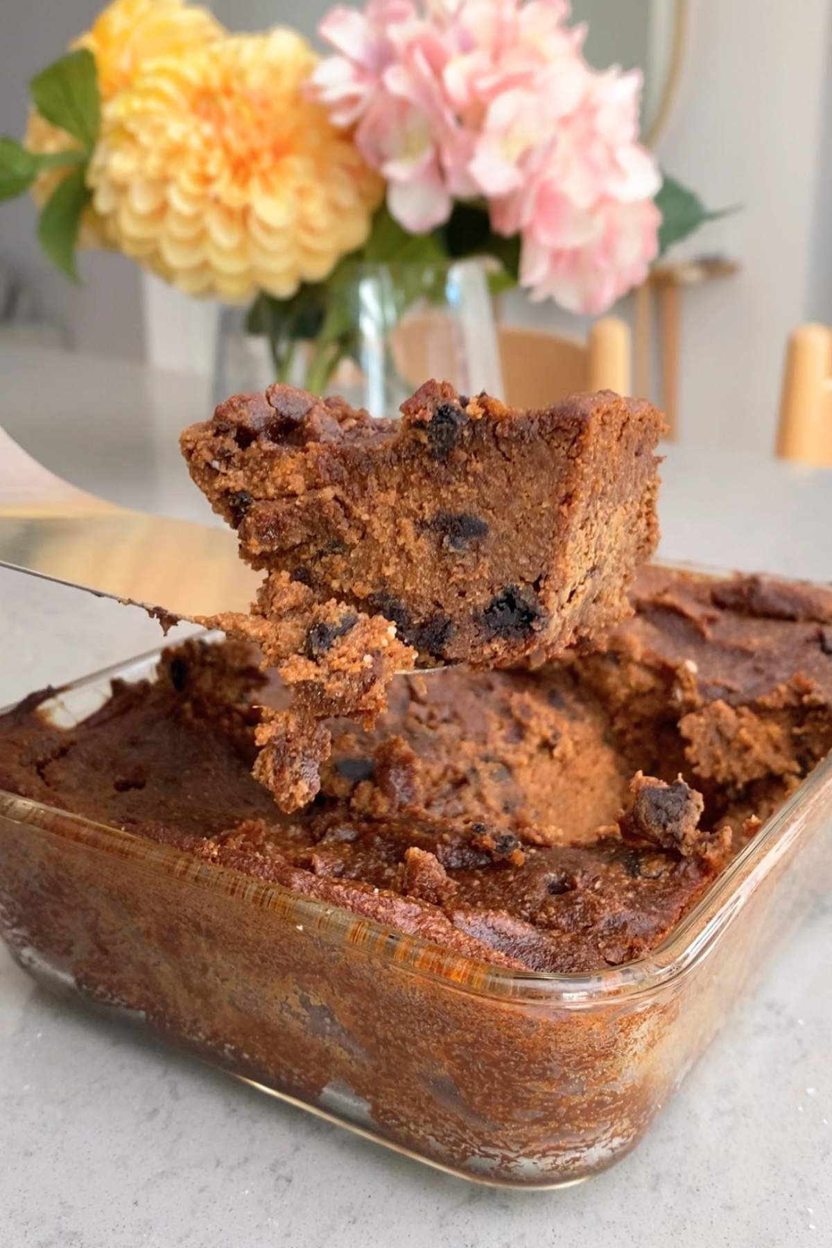A thick, gooey pumpkin brownie square with chocolate chips being lifted from a glass baking dish, with flowers blurred in the background.