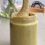 Viscous green spread drips from a spoon into a glass jar of butter.