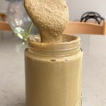 A spoon lifting creamy sunflower seed butter above a glass jar.
