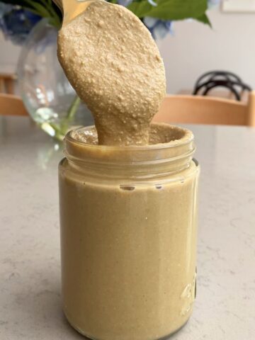 A spoon lifting creamy sunflower seed butter above a glass jar.