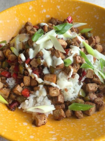 Crispy tofu cubes and diced peppers piled on white rice in a yellow bowl, drizzled with creamy mayo and scattered green onion slices.