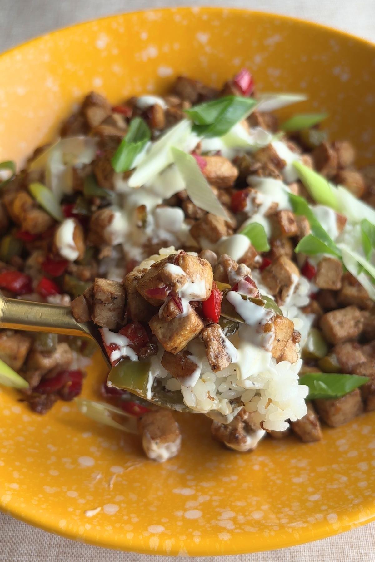 A spoonful of tofu sisig and rice lifted toward the camera, showing mayo drizzle and red and green pepper bits.