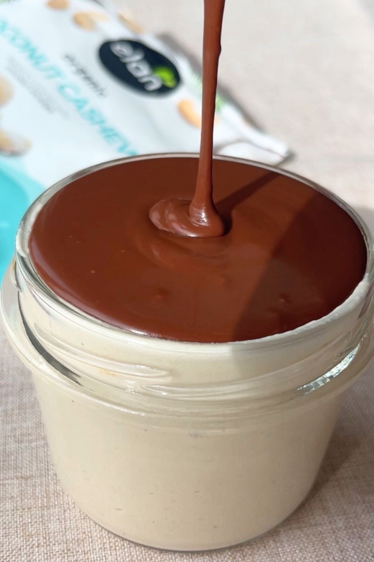 Smooth chocolate ganache being poured over creamy cashew mousse in a glass jar.