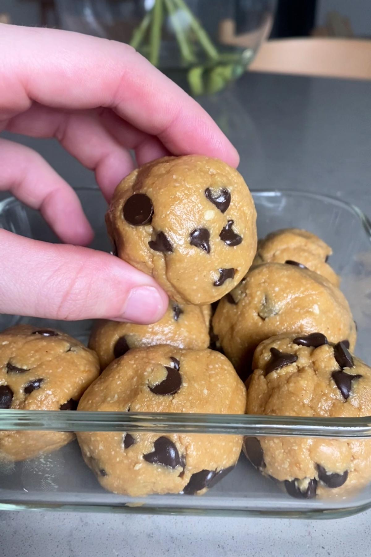 Hand holding a ball of dough with chocolate chips over a glass container filled with similar balls.