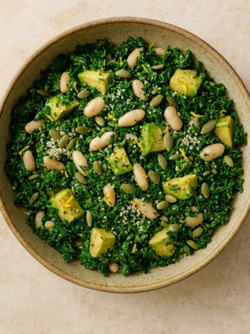 Fresh kale salad with avocado chunks, white beans, and seeds in a neutral ceramic bowl.