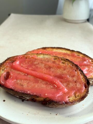 Slices of toasted bread spread with glossy pink guava jam, held in hand.