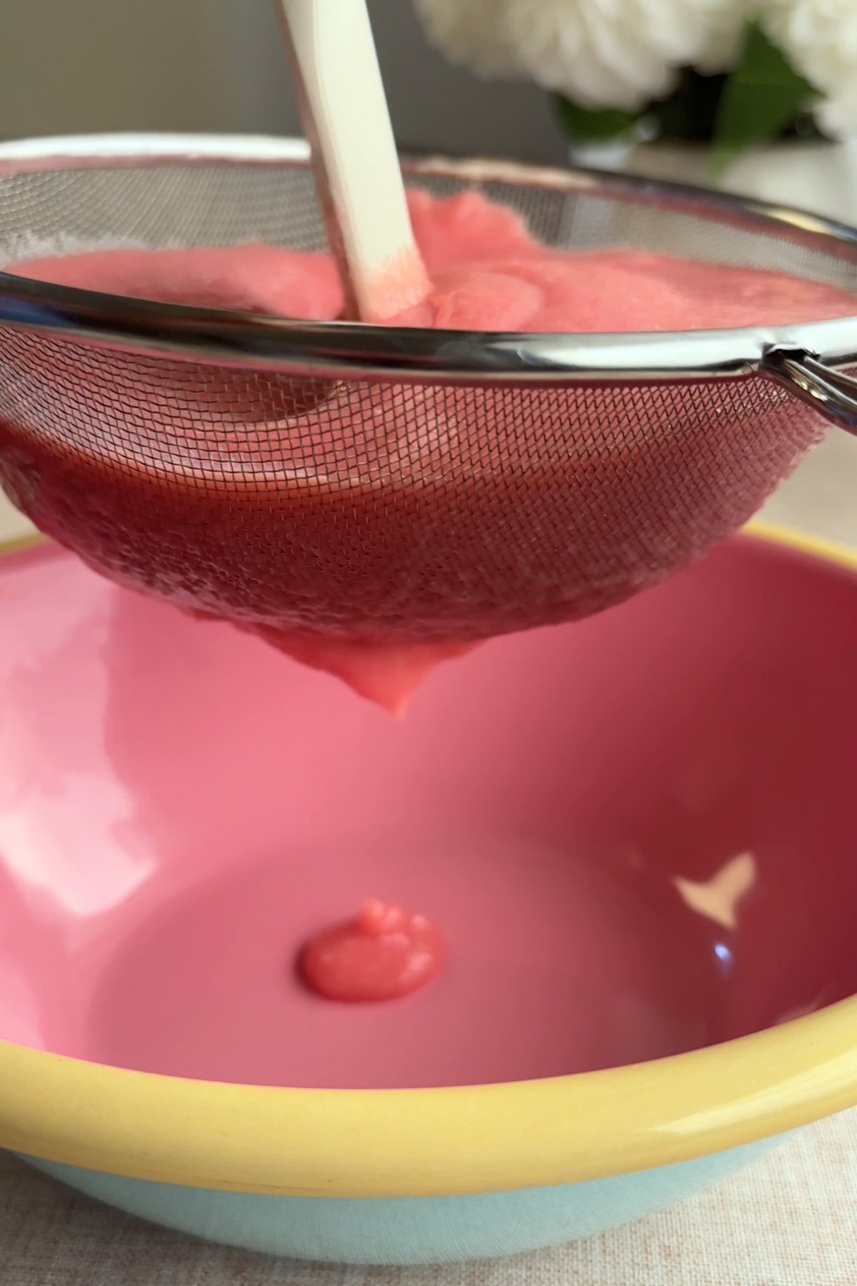 Pink guava puree being strained through a fine mesh sieve into a pink mixing bowl.
