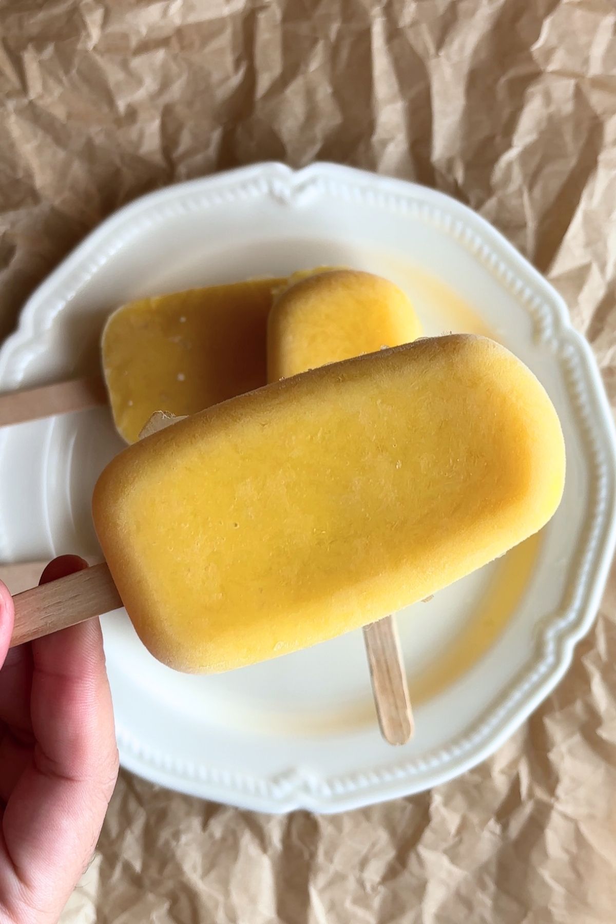 Frozen mango popsicle being held over a plate with another popsicle in the background.