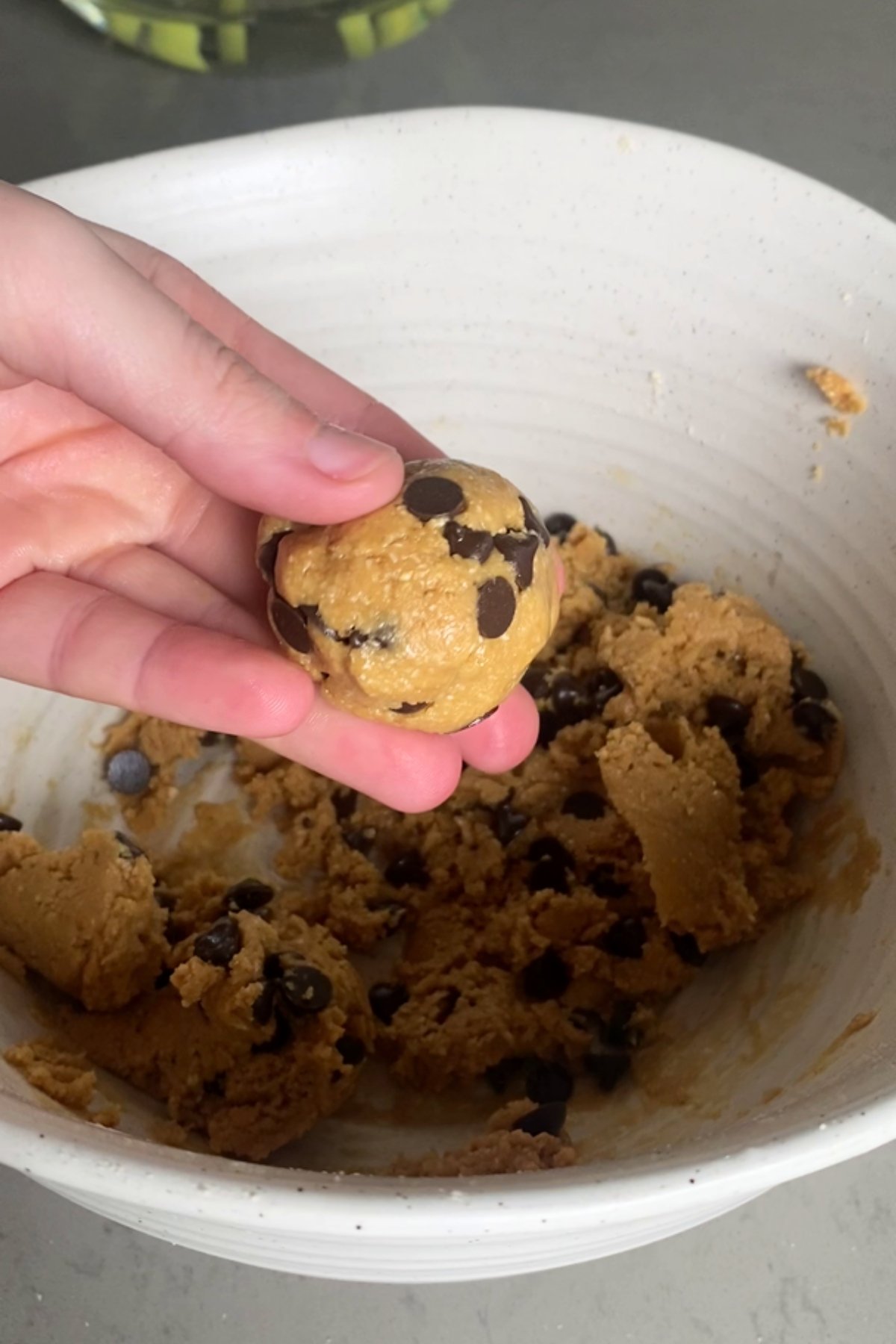 Hand holding a single dough ball with chocolate chips over a bowl of remaining dough.