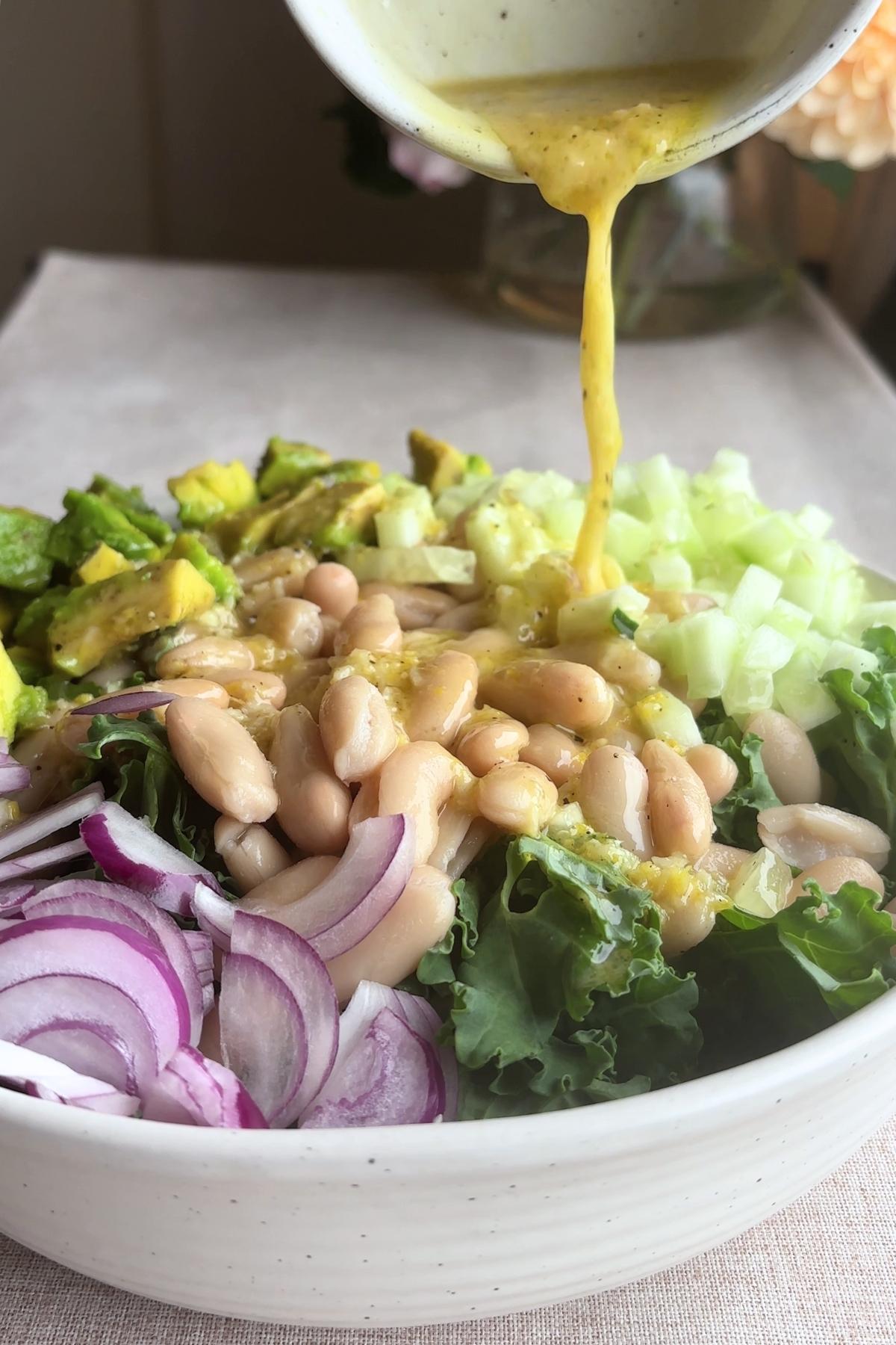 Dressing being poured over kale, beans, avocado, and cucumber.