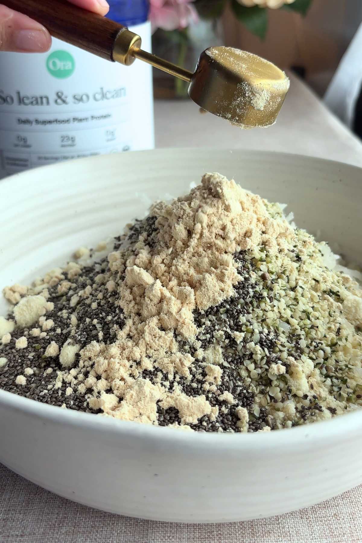 Measuring scoop of protein powder over chia seeds and hemp seeds in a mixing bowl.