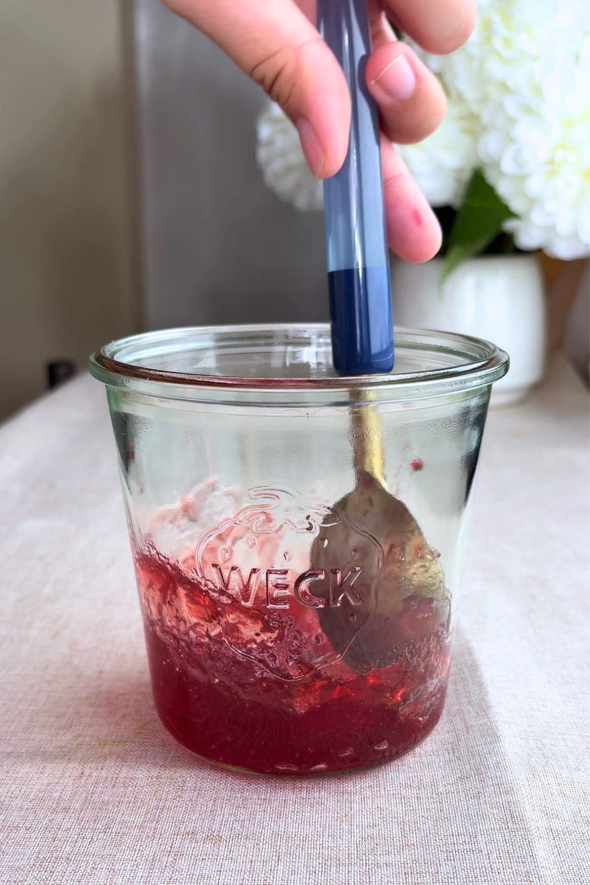 Spoon stirring chunky red strawberry jam in a clear glass jar.
