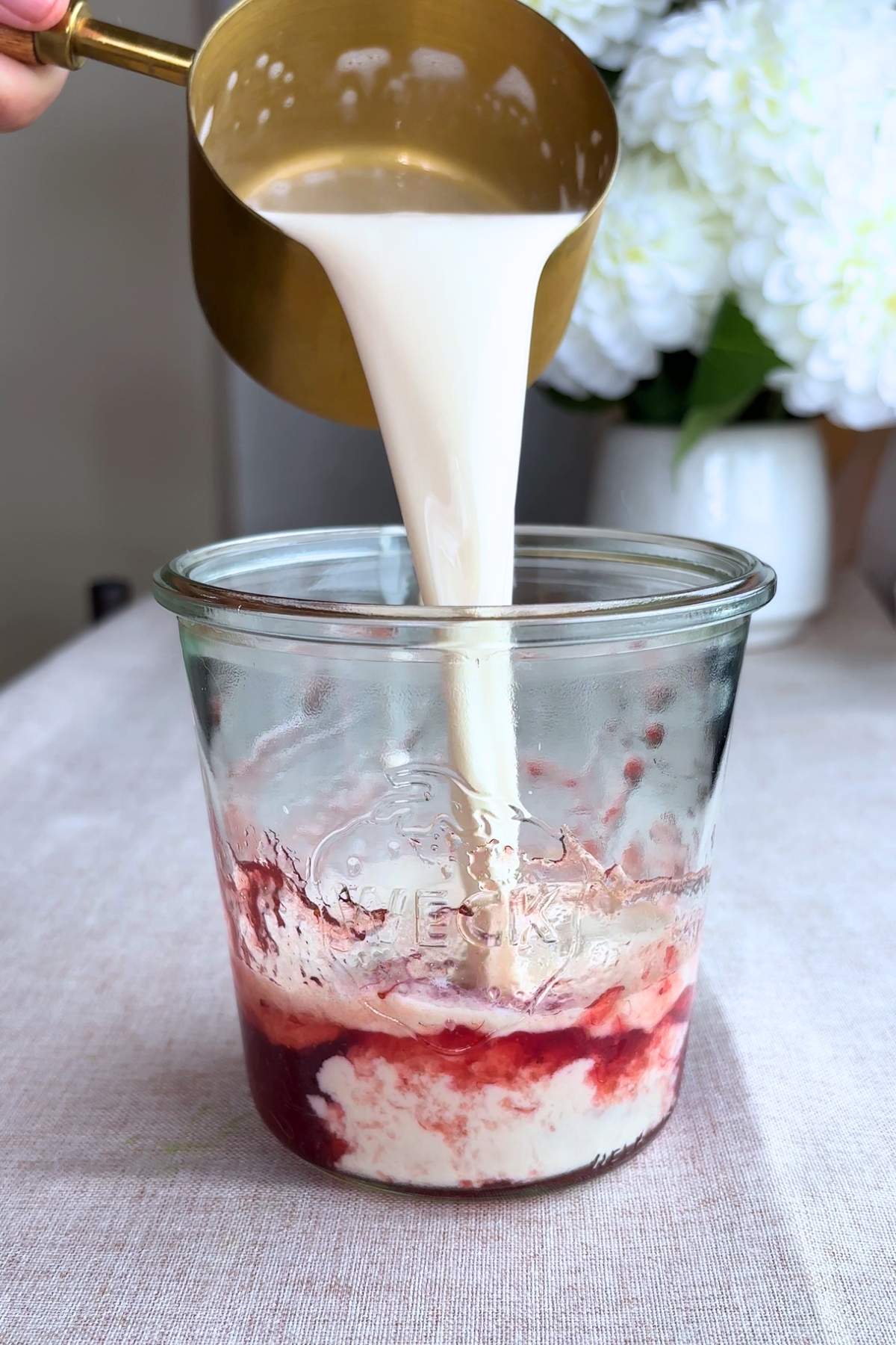 Milk being poured from a gold measuring cup into a glass with strawberry jam.