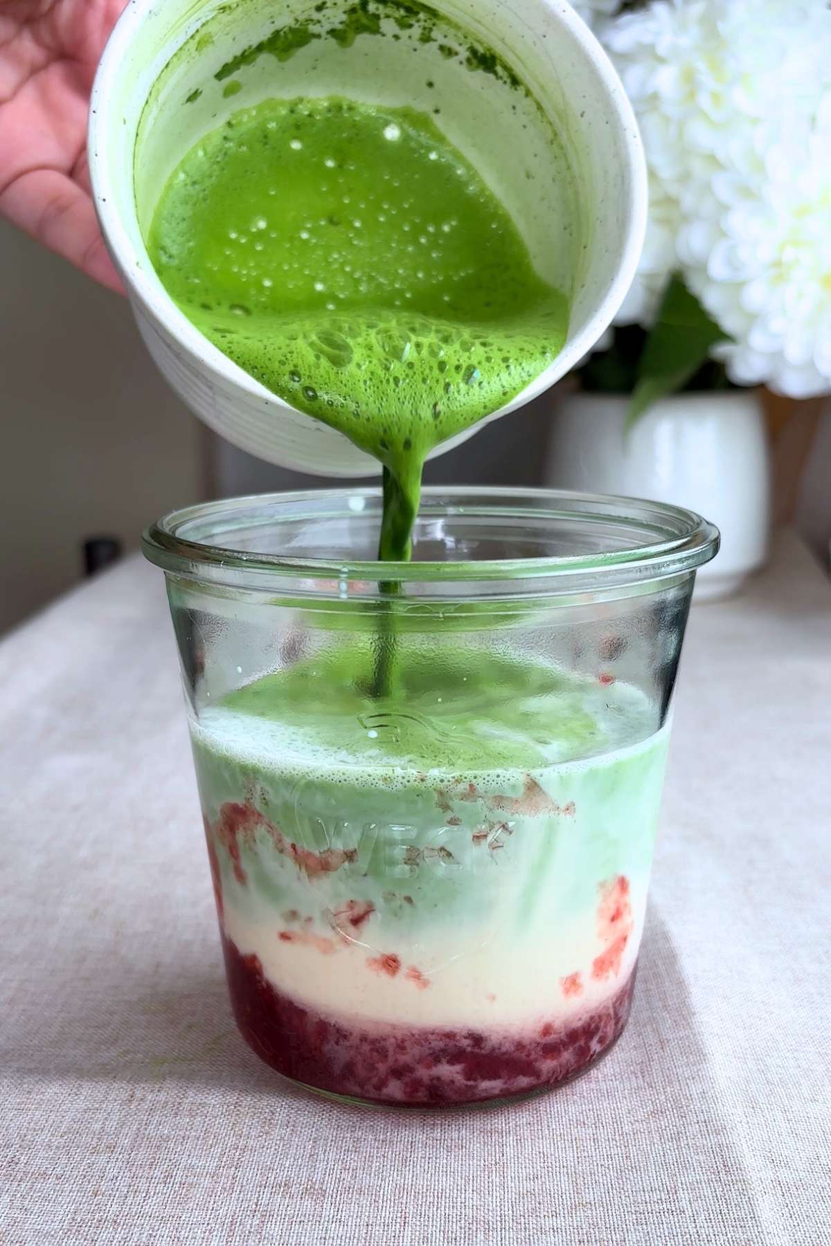 Smooth green matcha being poured over milk and strawberries in a glass jar.
