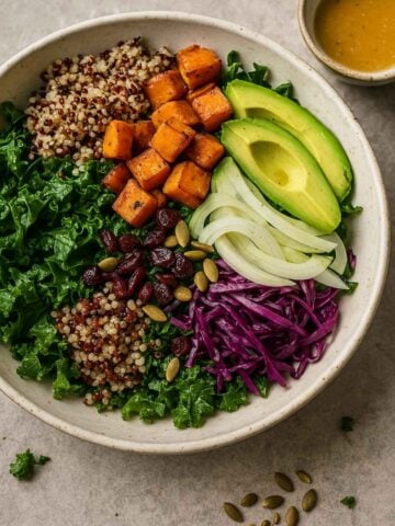 Roasted sweet potato, quinoa, avocado, kale, cabbage, onion, cranberries, and pumpkin seeds arranged in a bowl.