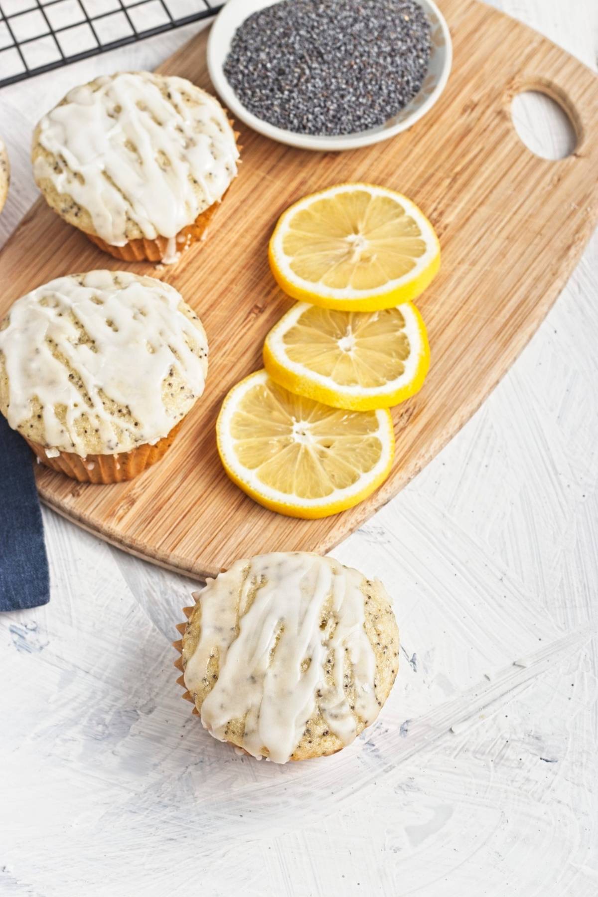 Glazed muffins on a wooden board with lemon slices and a dish of poppy seeds.