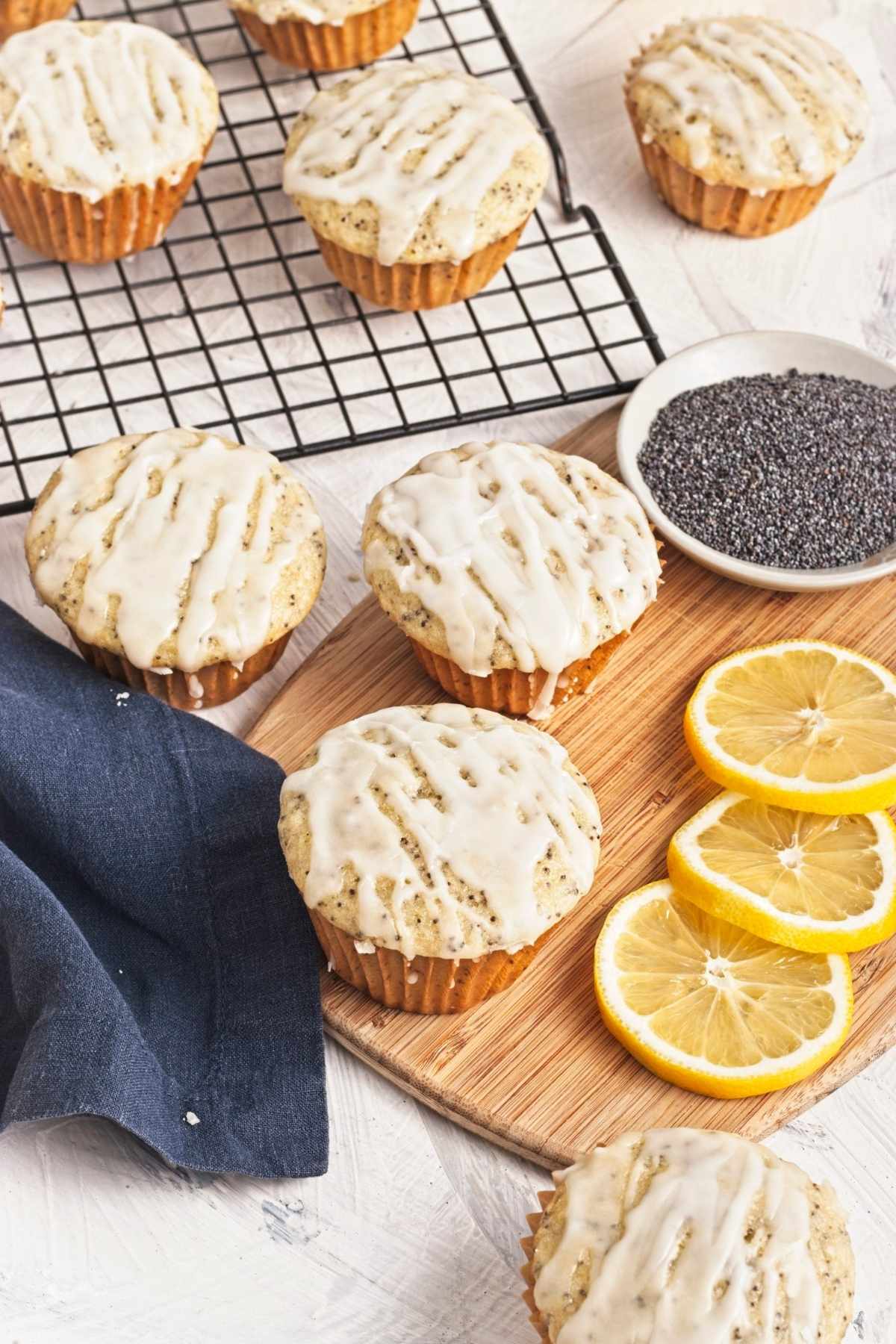 Glazed muffins on a wooden board with lemon slices and a dish of poppy seeds.