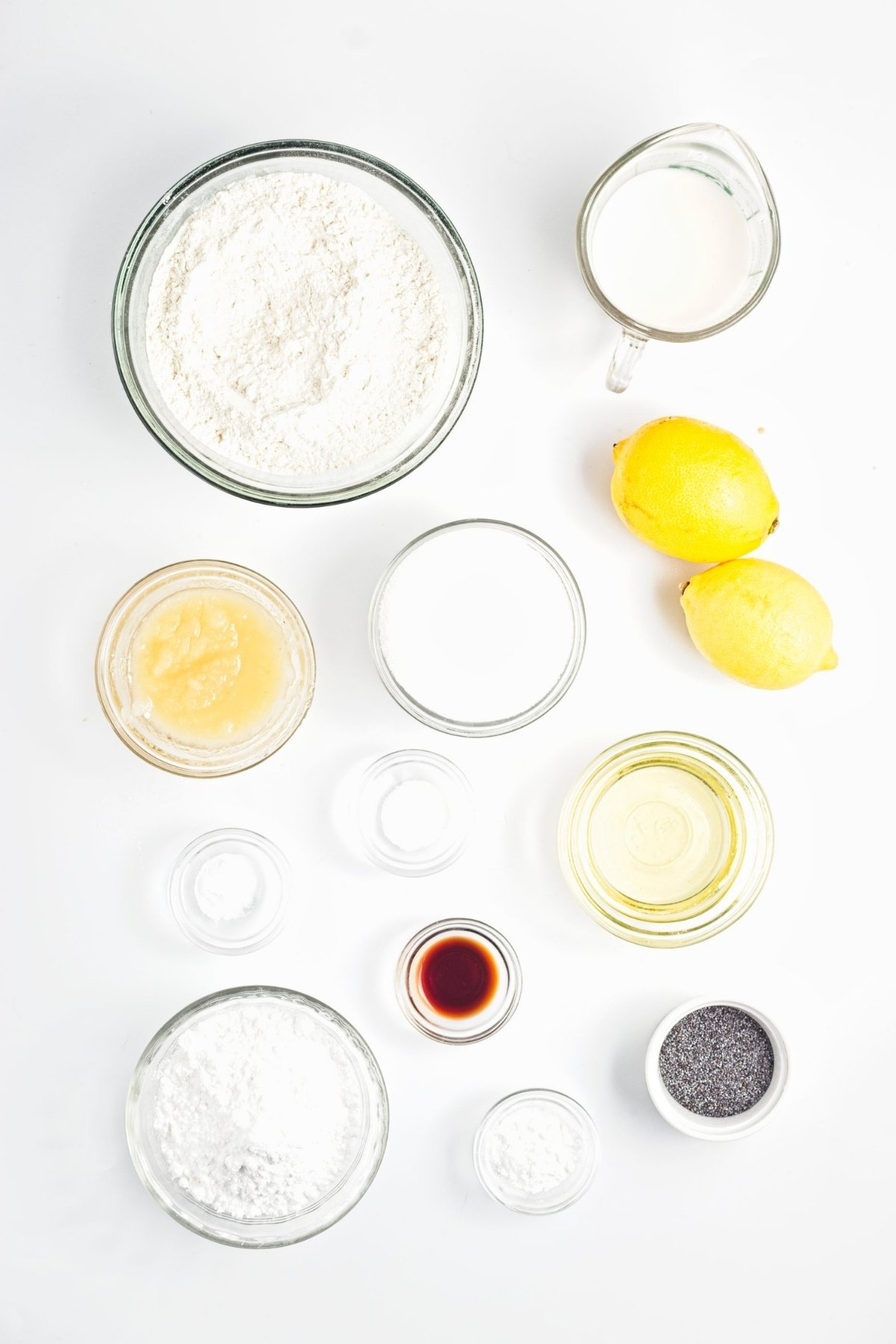 Measured flour, sugar, applesauce, milk, lemons, oil, poppy seeds, and other baking items arranged on white surface.