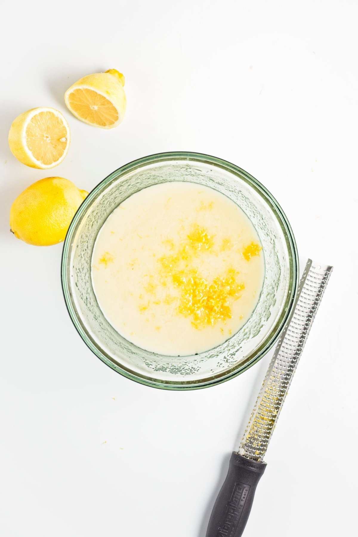 Bowl of wet mixture with lemon zest sprinkled on top, zester and halved lemons nearby.