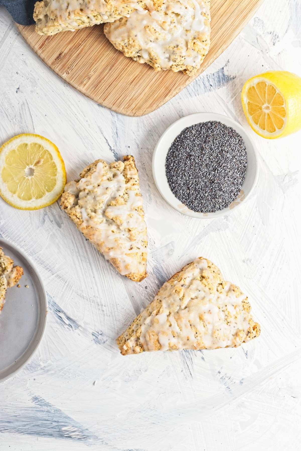 Glazed triangular scones on a wooden board with lemon slices and a dish of poppy seeds.