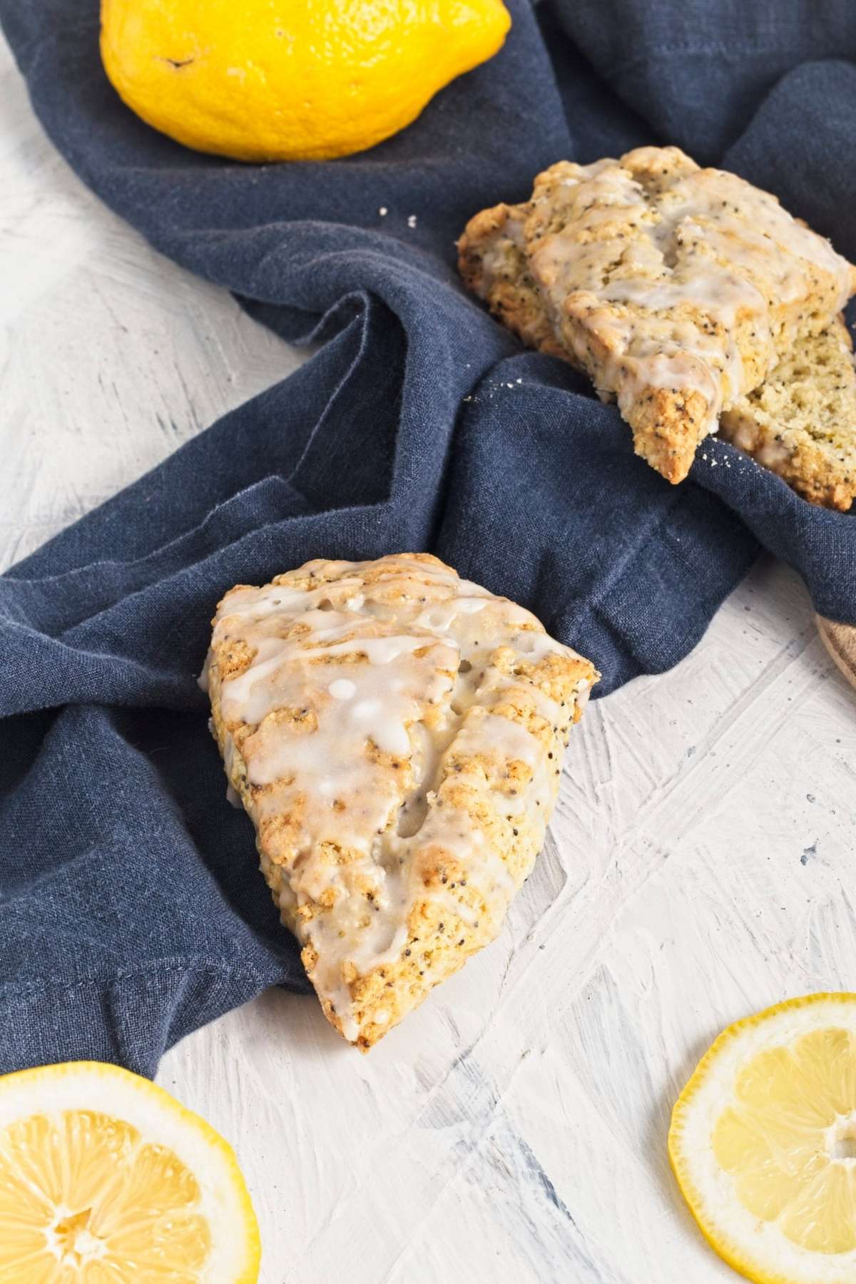 Glazed scone resting on a blue cloth, with a whole lemon and lemon slices nearby.