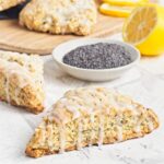 Close-up of a glazed scone in front of a dish of poppy seeds, with more scones and lemon slices in the background.