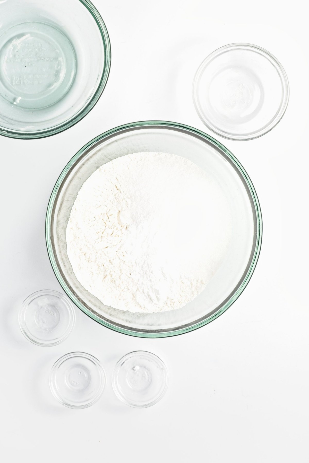Bowl of flour and dry baking ingredients in glass bowls on a white surface.