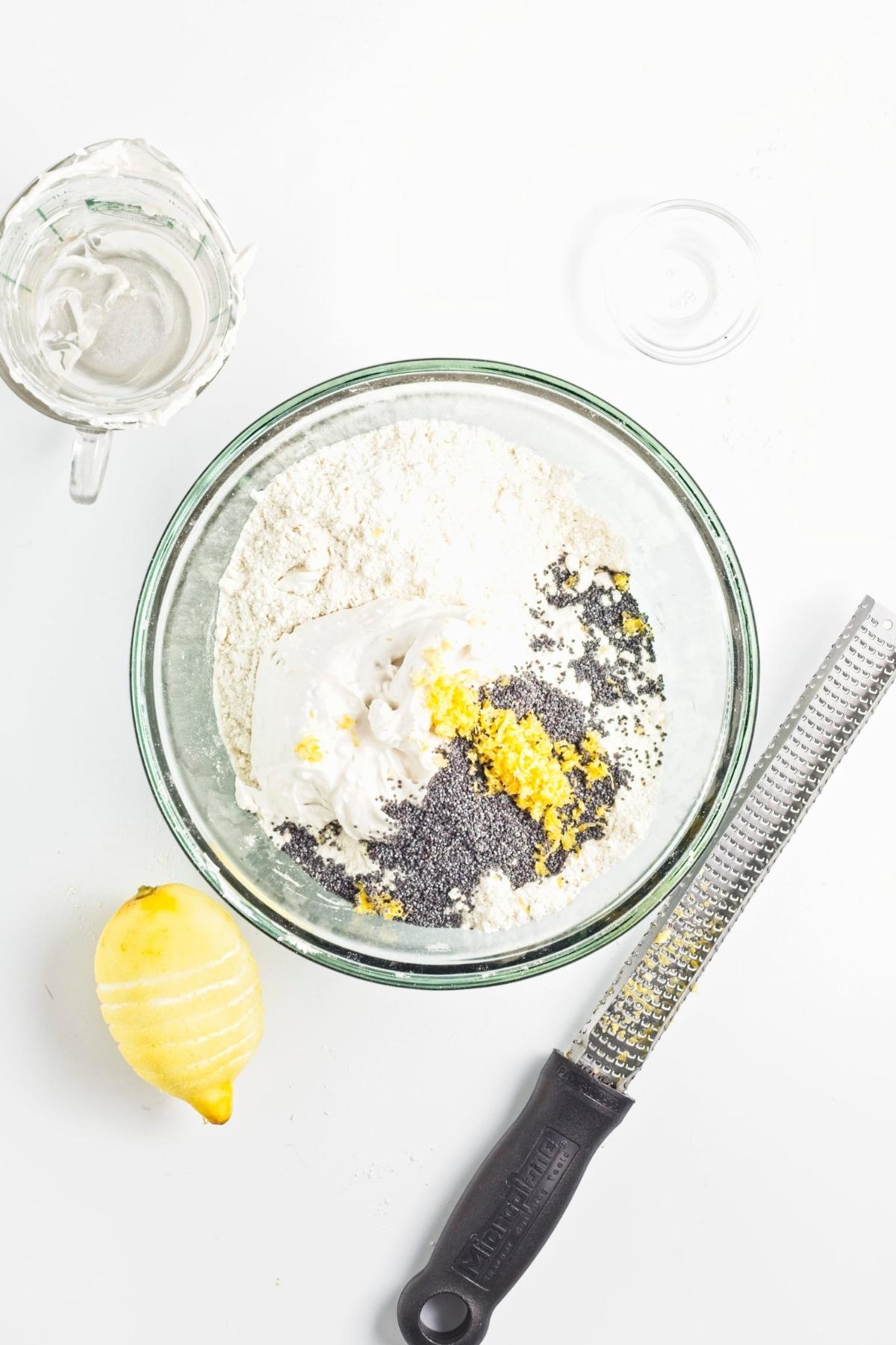Mixing bowl with flour, coconut cream, lemon zest, and poppy seeds, with a lemon and zester nearby.