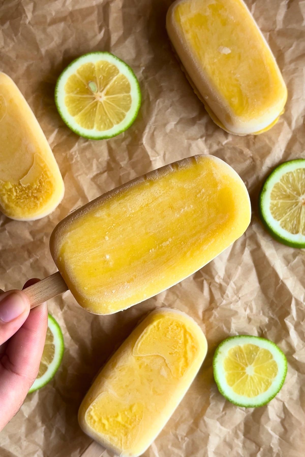 Hand holding a frozen mango popsicle on a stick with lime slices scattered around.