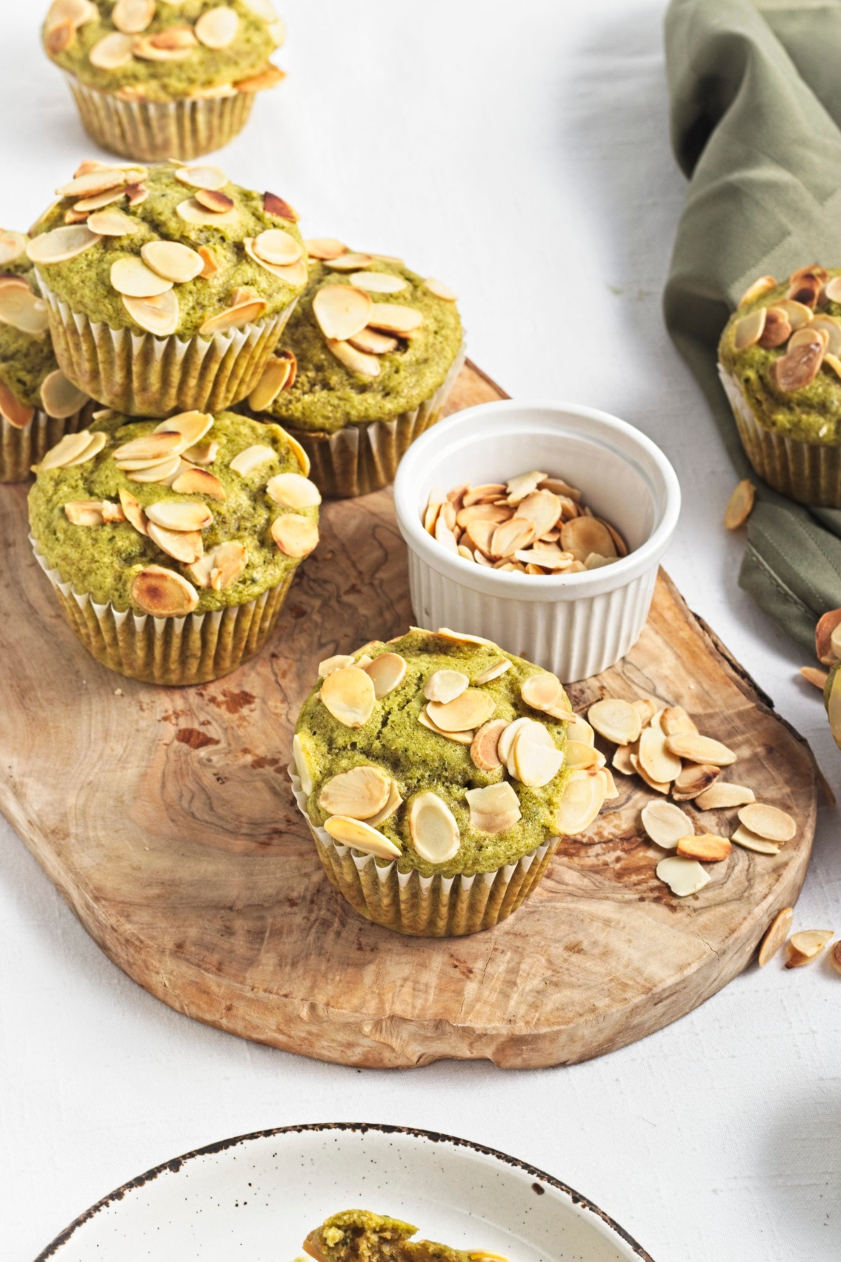 Green muffins topped with toasted almond slices on a wooden board beside a small bowl of almonds.