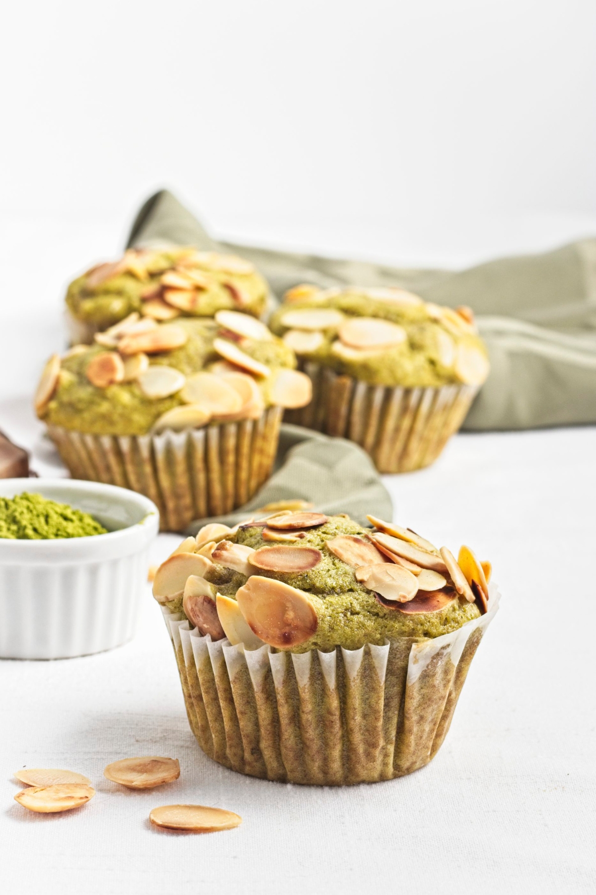 Green muffin with toasted almond topping in focus, with more muffins and a bowl of matcha powder behind.