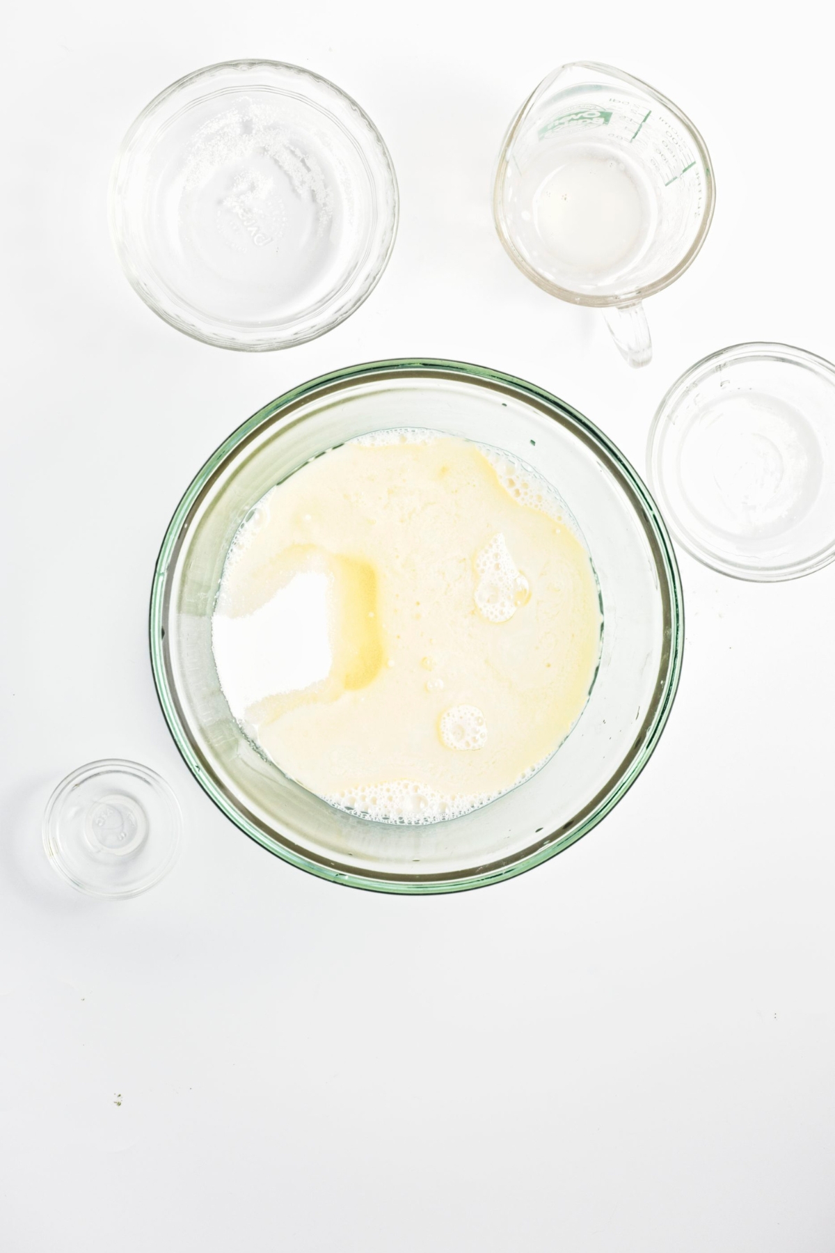 Large glass bowl with almond milk, oil, vinegar, and sugar mixture surrounded by smaller bowls.