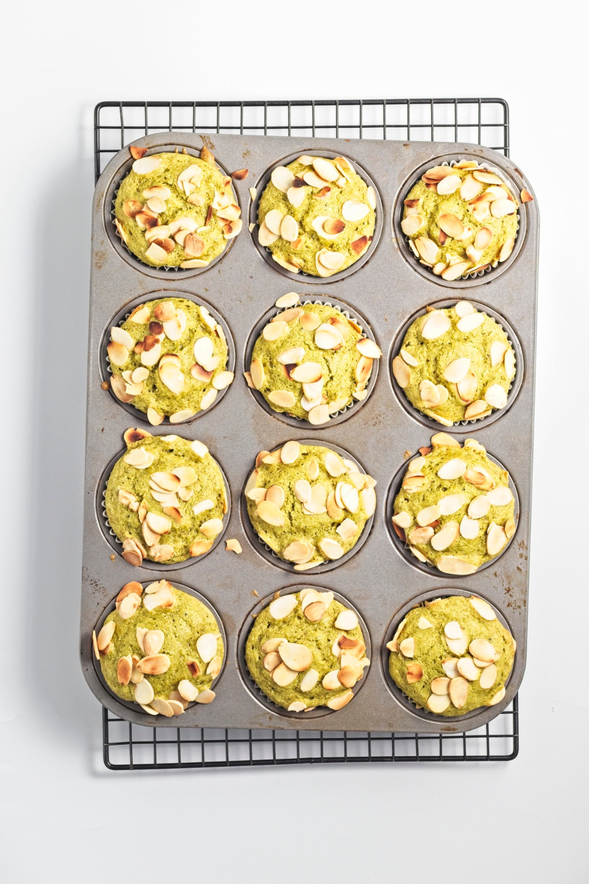 Baked green muffins with toasted almond tops in a muffin tin on a cooling rack.