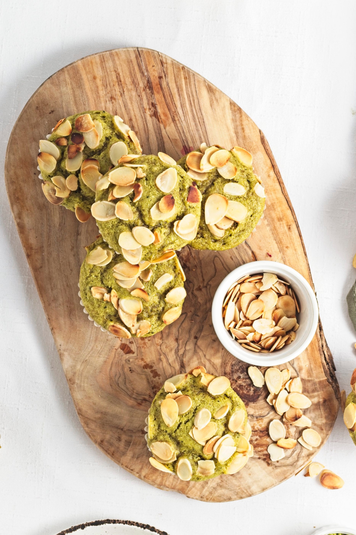 Green muffins topped with toasted almonds on a wooden board next to a small bowl of almonds.