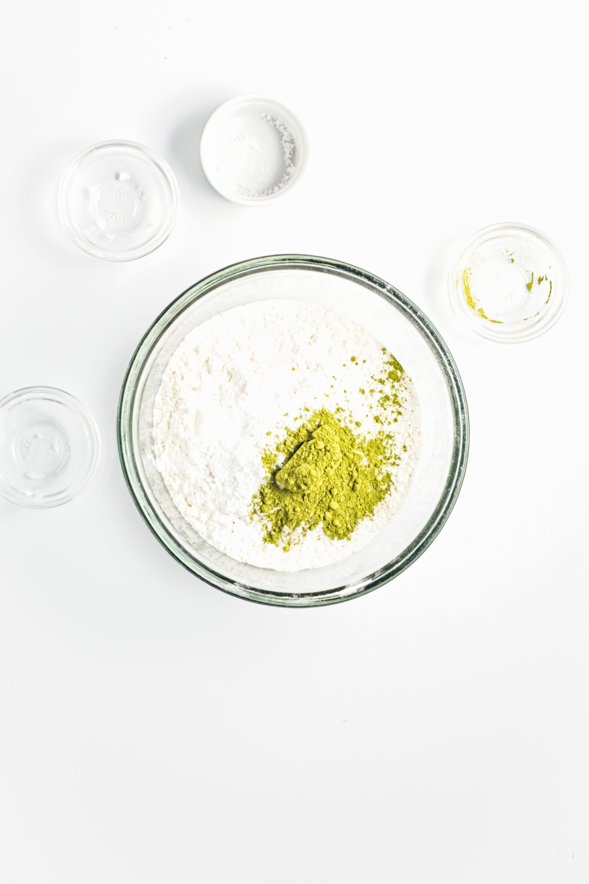 Flour mixture in a glass bowl with matcha powder on top, surrounded by empty bowls.