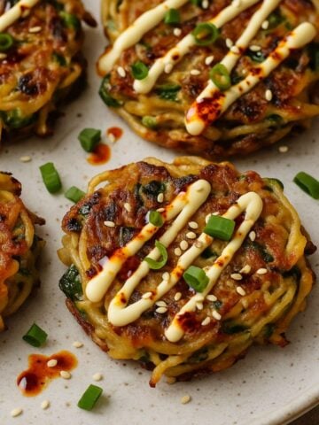 Crispy golden pancakes topped with mayo drizzle, chili oil, sesame seeds, and chopped green onions on a plate.