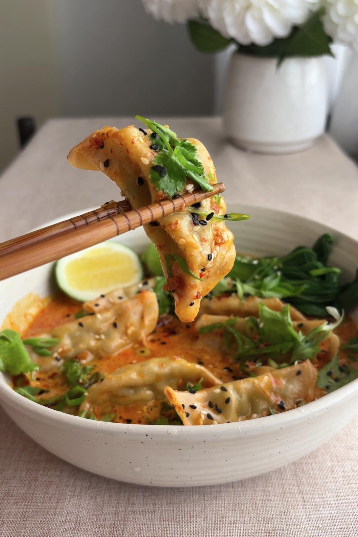 Dumpling lifted with chopsticks over curry soup, garnished with sesame seeds and herbs.