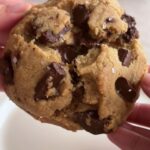Hands holding a thick cookie with visible melted chocolate chunks.