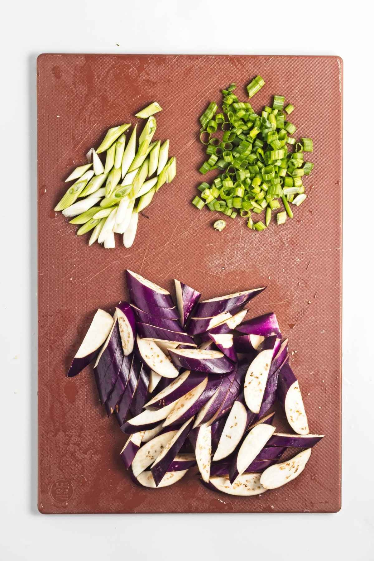 Sliced eggplant and green onions on a brown cutting board, separated into white/light and dark green portions.