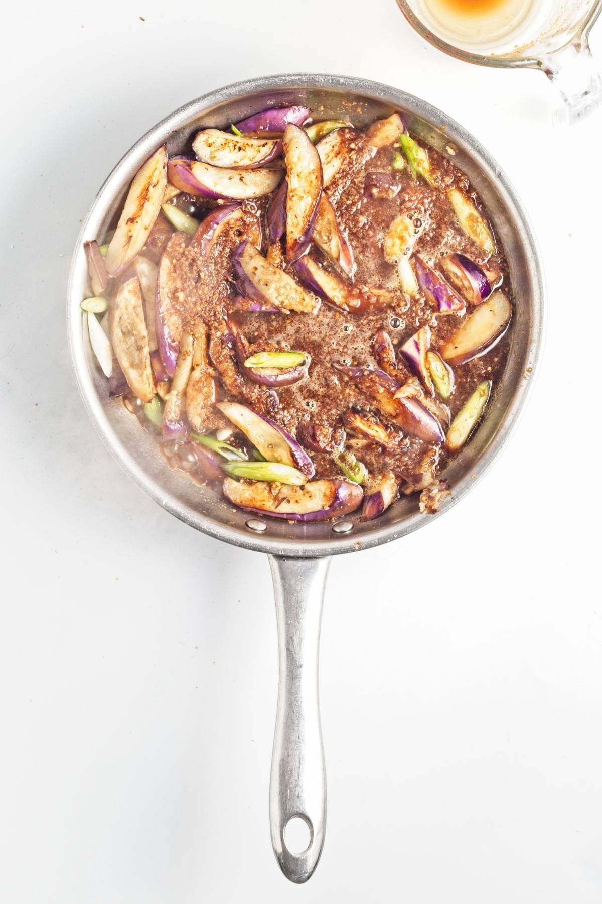 Eggplant slices and green onions simmering in sauce in a stainless steel pan.