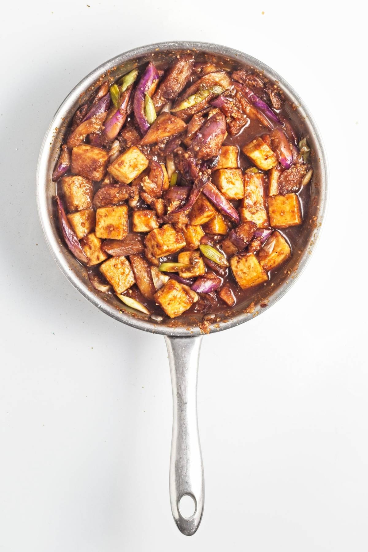 Tofu cubes, eggplant, and green onions coated in a thick brown sauce in a stainless steel pan.
