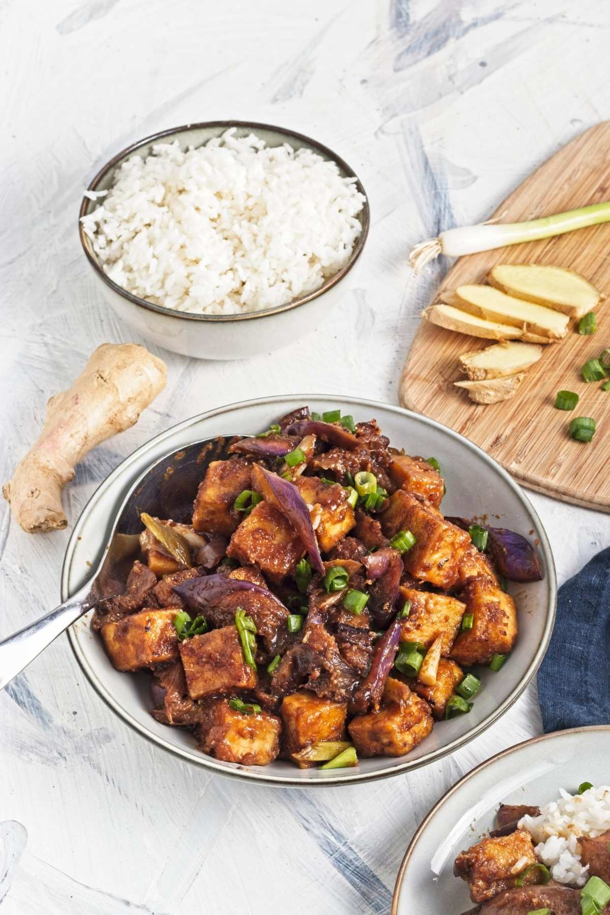 Bowl of tofu and eggplant with green onions, with rice and sliced ginger on a board in the background.