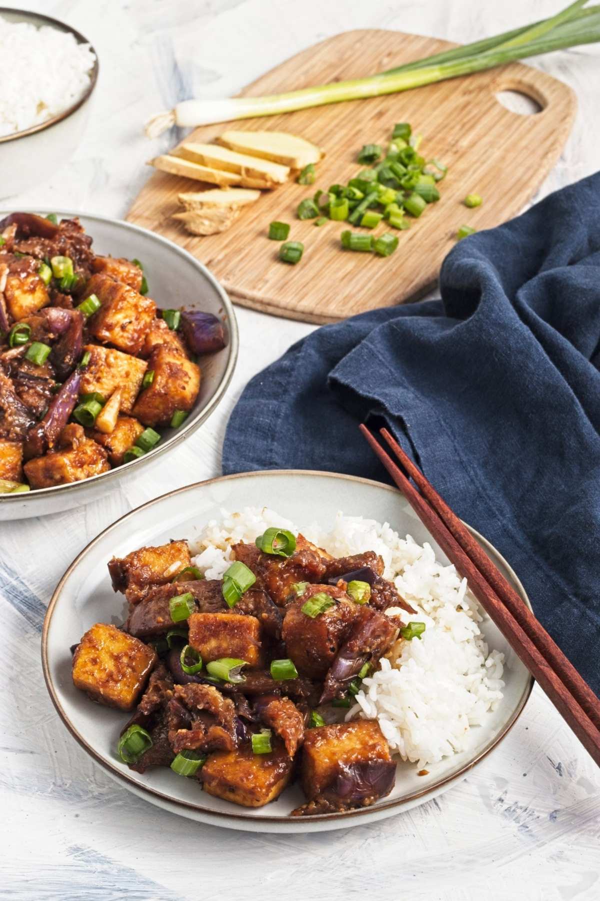 Plates of tofu and eggplant with rice, with a board of sliced ginger and green onions in the background.