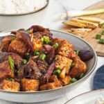 Close-up of tofu and eggplant in sauce, garnished with green onions, with rice and ginger in the background.