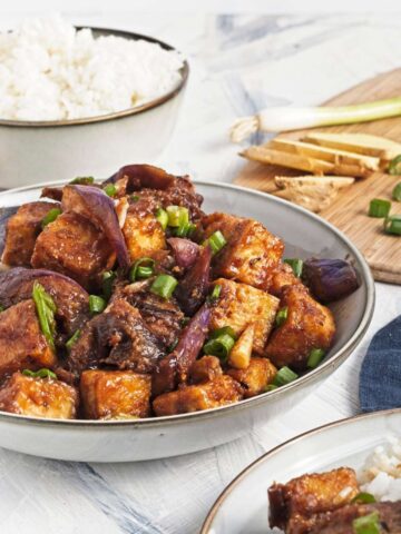 Close-up of tofu and eggplant in sauce, garnished with green onions, with rice and ginger in the background.