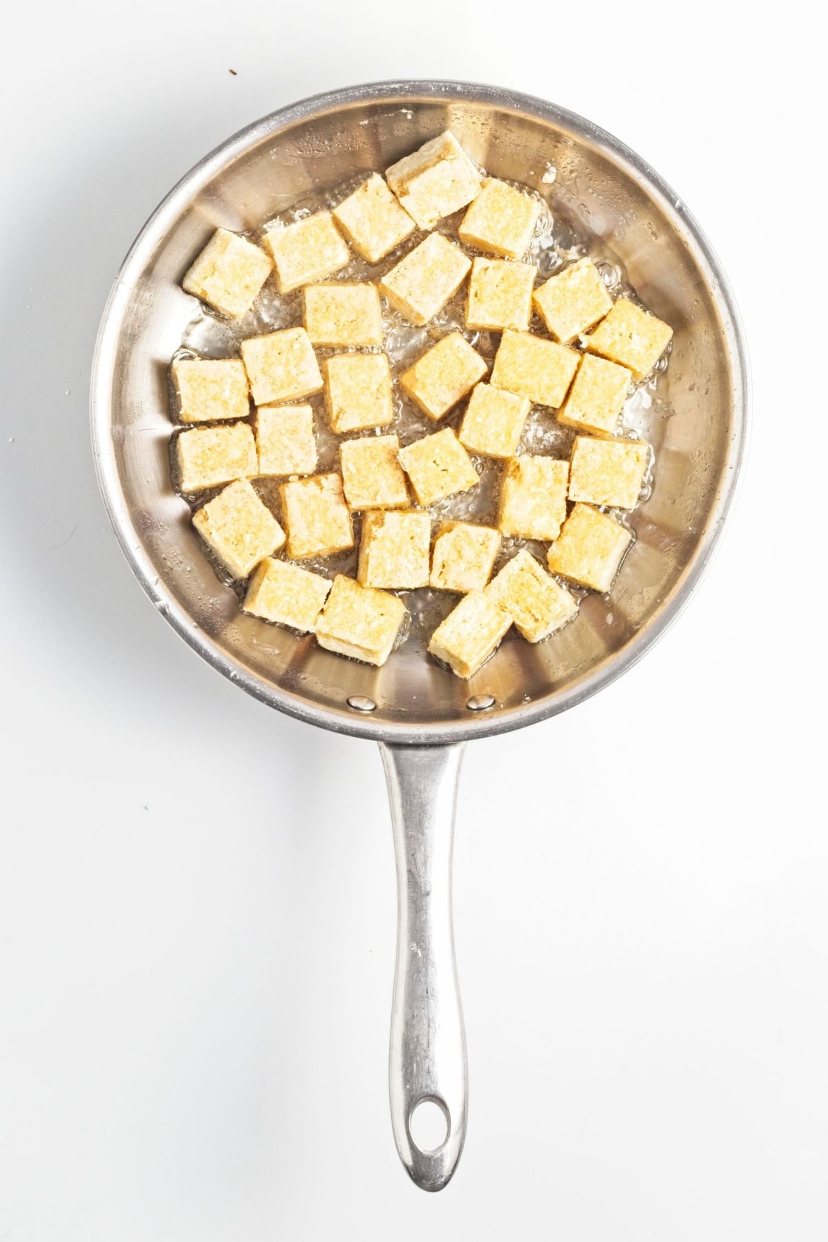 Cubes of tofu coated in cornstarch frying in oil in a stainless steel pan.