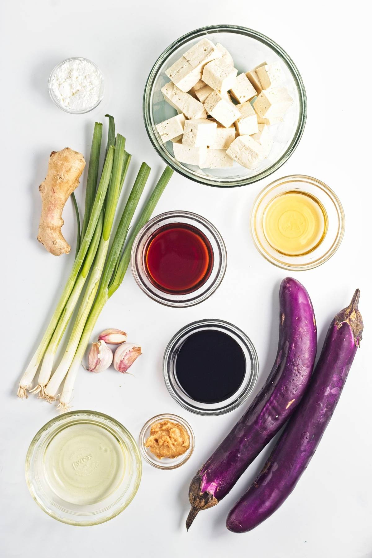 Cubes of tofu, long eggplants, green onions, ginger, garlic, sauces, oil, and cornstarch arranged on a white surface.